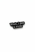 a black enamel pin with white text that says wait i see a rock on a white background