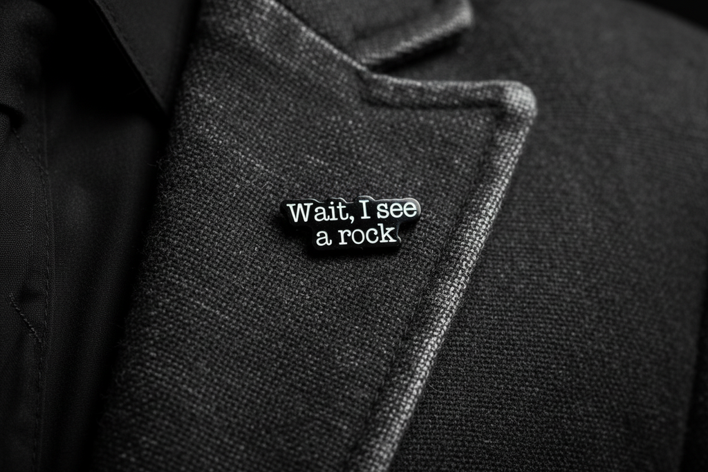 a black enamel pin with white text that says wait i see a rock on a gray lapel background