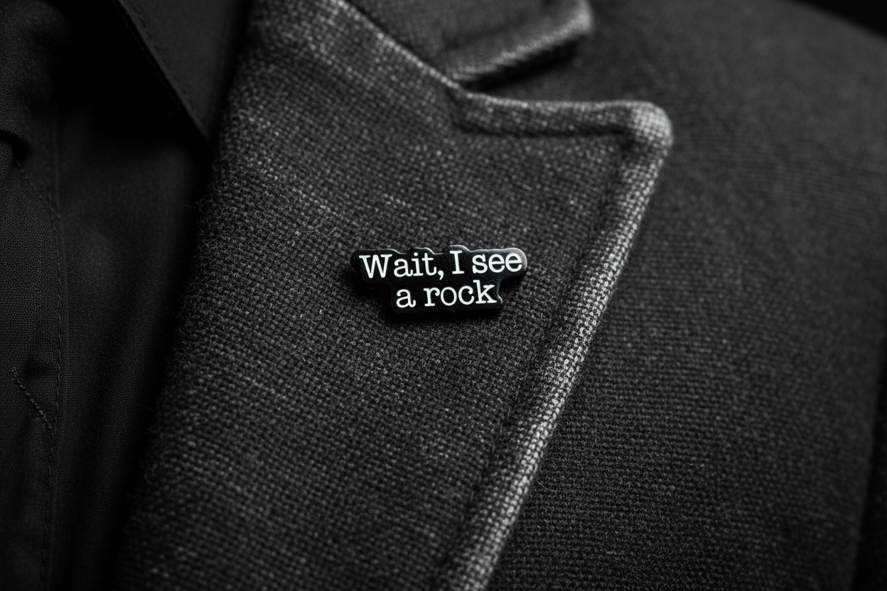 a black enamel pin with white text that says wait i see a rock on a gray lapel background