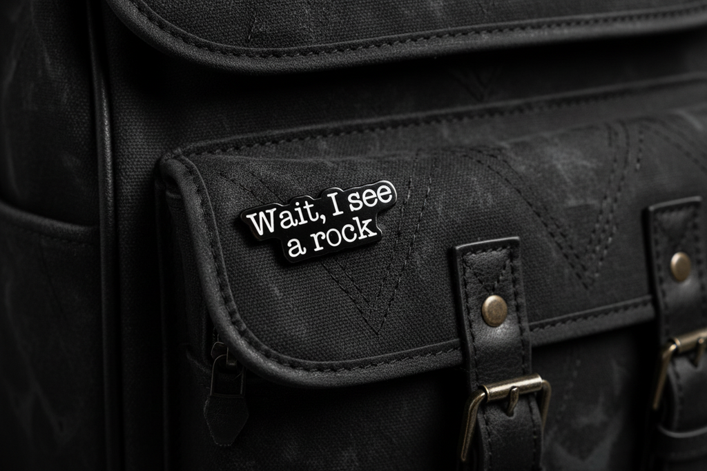 a black enamel pin with white text that says wait i see a rock on a black bookbag background