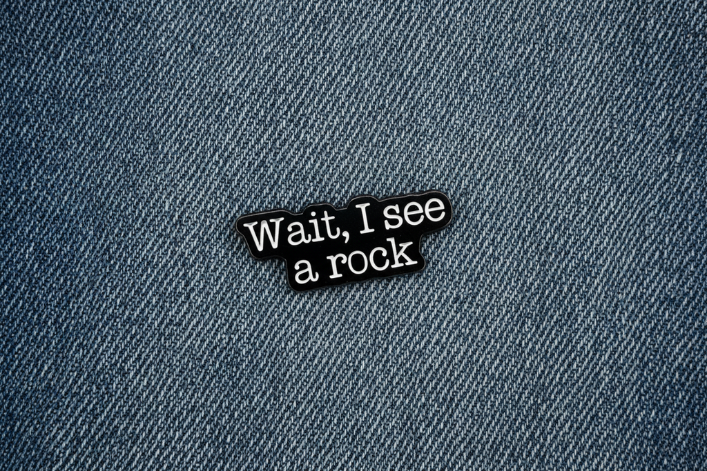 a black enamel pin with white text that says wait i see a rock on a denim background