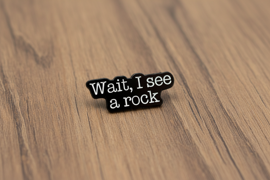 a black enamel pin with white text that says wait i see a rock on a wood background