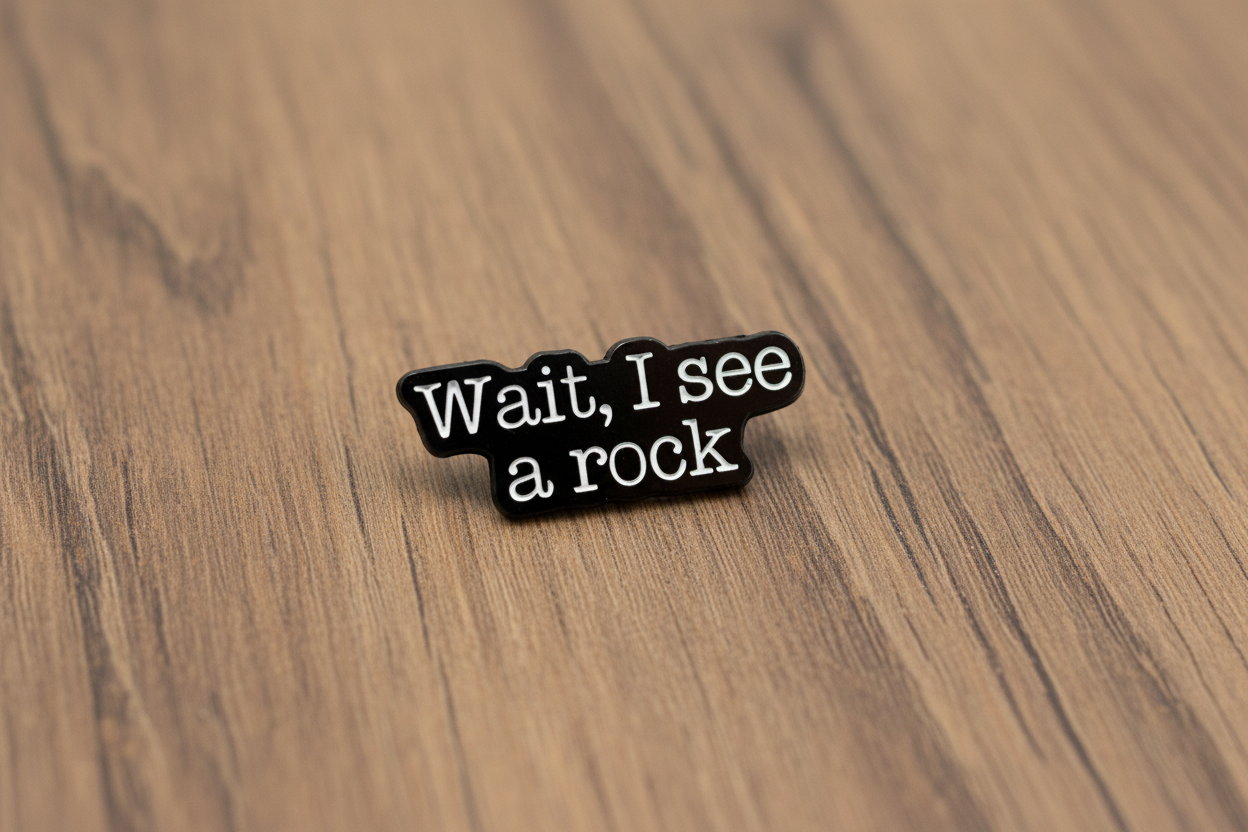 a black enamel pin with white text that says wait i see a rock on a wood background