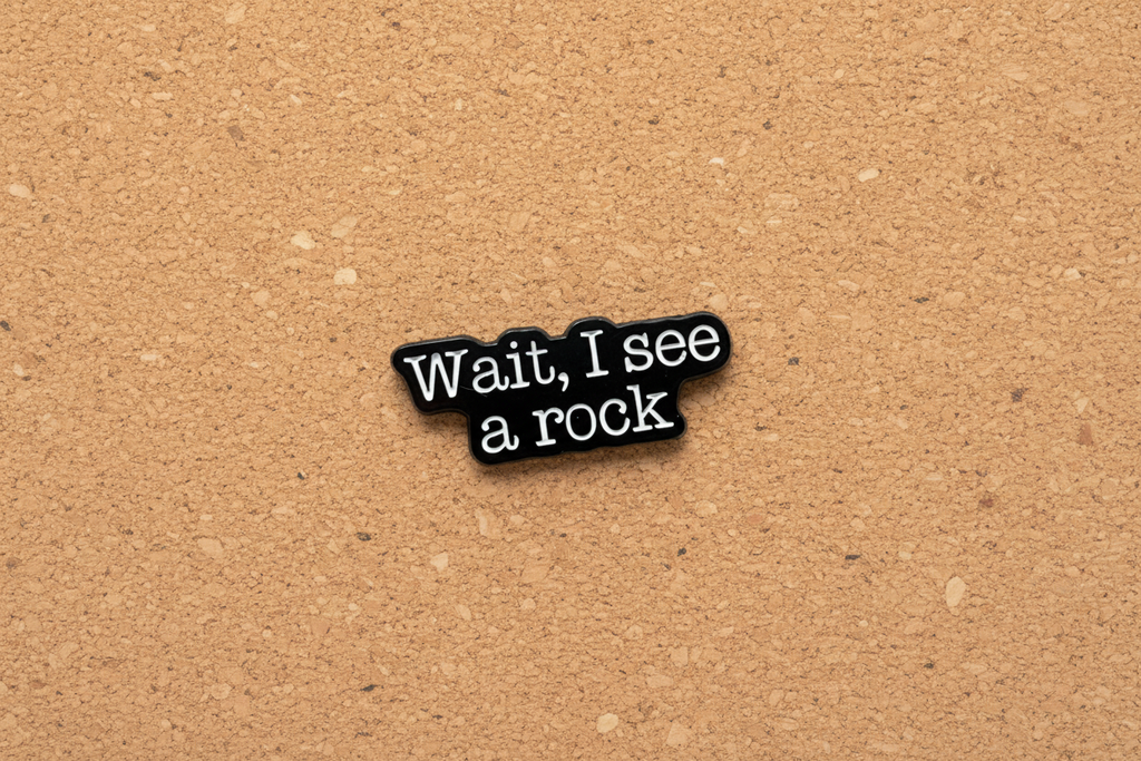 a black enamel pin with white text that says wait i see a rock on a cork background