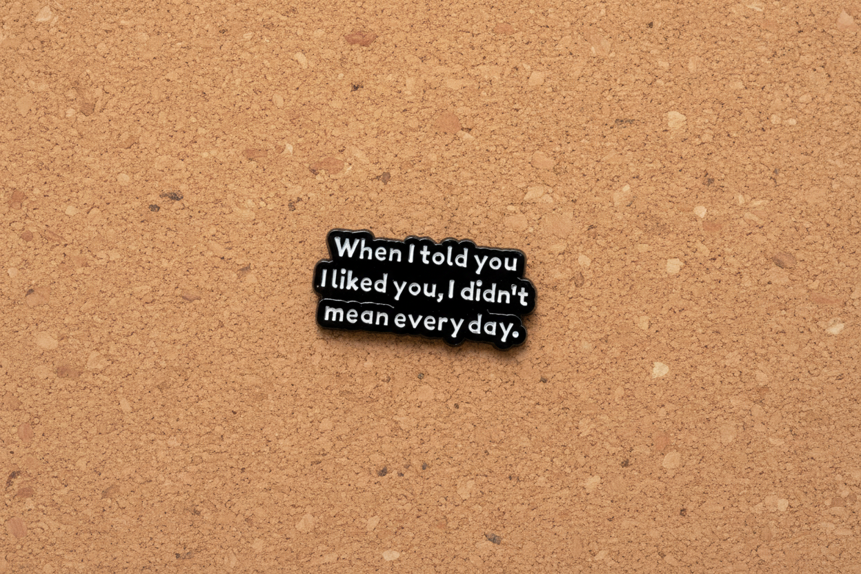 black enamel pin with white text and a sarcastic saying on a cork background
